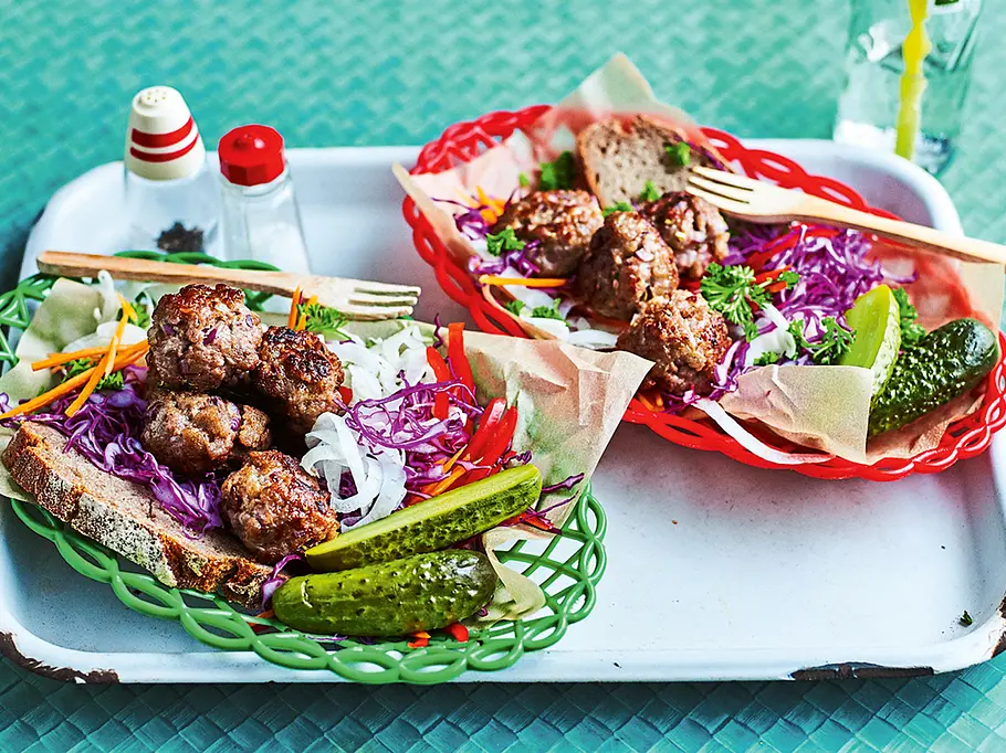 Buntes Brotzeit-Körbchen Auf einem weißen Emaille-Tablett stehen zwei bunte Kunststoffkörbchen mit einer Scheibe Bauernbrot, auf der ein bunter Rohkostsalat aus Rotkohl, Möhren, Fenchel und Paprika unter mehreren kleinen Hackbällchen und angeschnittener Cornichons liegt.