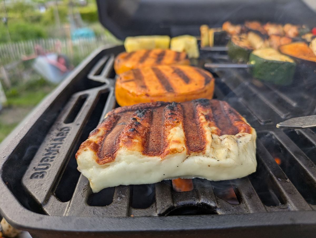 burnhard-gasgrill-wayne-grillrost Nahaufnahme: Auf dem Burnhard Wayne Grillrost werden Grillkäse und Gemüse mit leckeren Grillstreifen zubereitet, im Hintergrund ist unscharf ein Garten zu erkennen.
