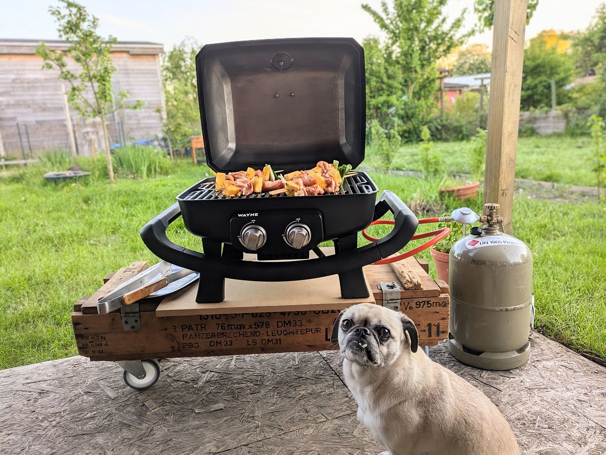 burnhard-gasgrill-wayne-polli Ein Burnhard Wayne Grill mit Spießen steht auf einem Tisch, daneben eine Gasflasche im Garten und im Vordergrund sitzt ein kleiner Hund.