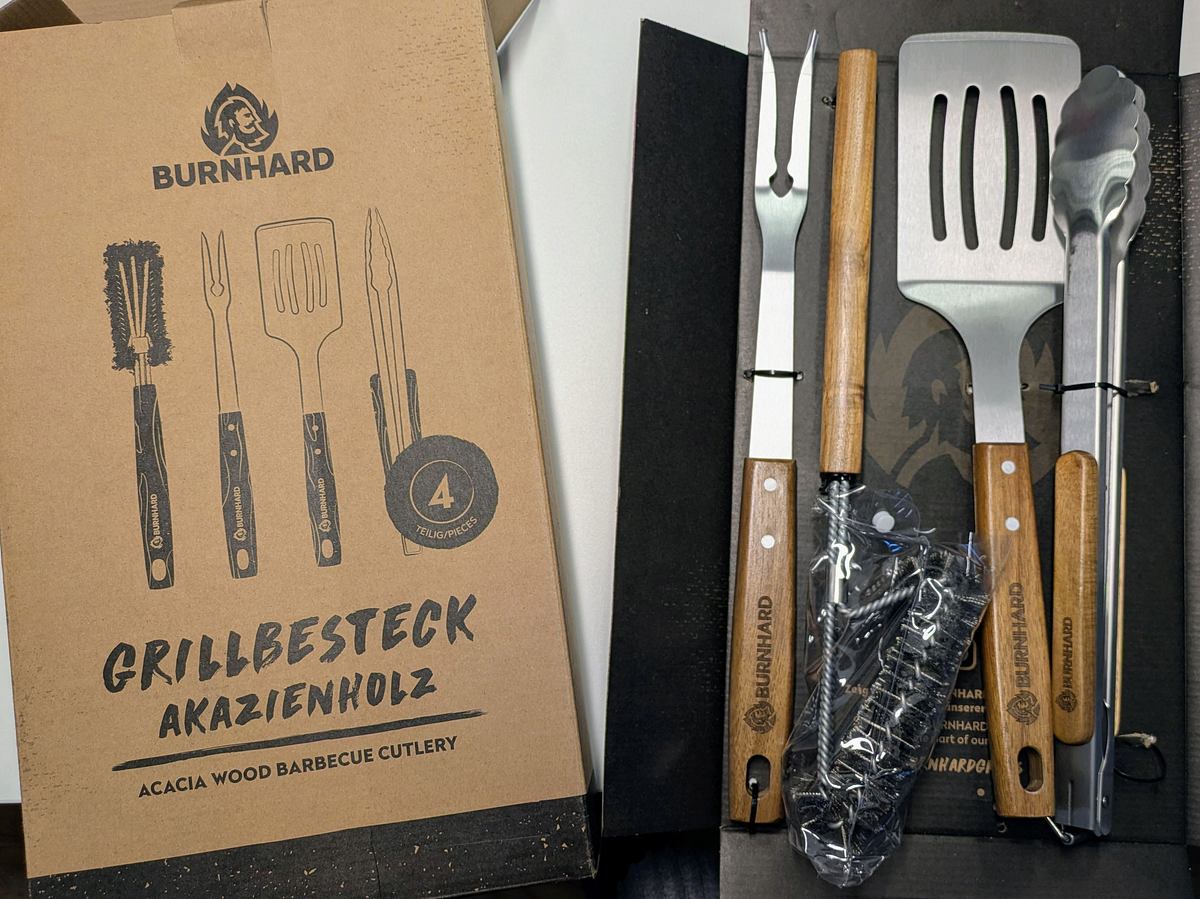 Vierteiliges Burnhard Grillbesteckset aus Edelstahl und Akazienholz liegt geöffnet neben der Verpackung auf einem Tisch.