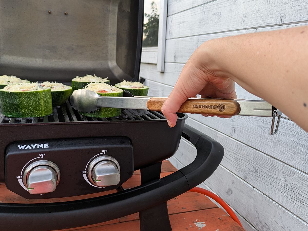 Eine Burnhard Grillzange hält ein gefülltes Zucchinistück auf dem Burnhard Wayne Gasgrill, der Deckel ist geöffnet.