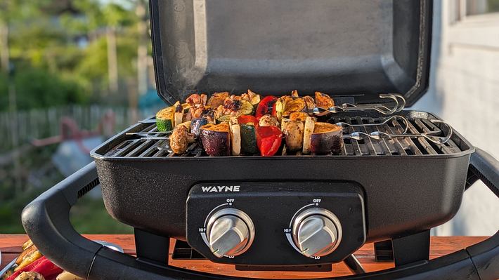 Der offene Gasgrill Wayne der Marke Burnhard ist zu sehen. Auf dem Rost liegen verschiedene Spieße mit buntem Gemüse und Fleisch. Vorne am Grill befinden sich zwei silberne Drehknöpfe. Im Hintergrund sieht man einen Garten mit Bäumen. Die Szene deutet auf einen sonnigen Grillabend hin. - Foto: LECKER.de/Emily Beckmann
