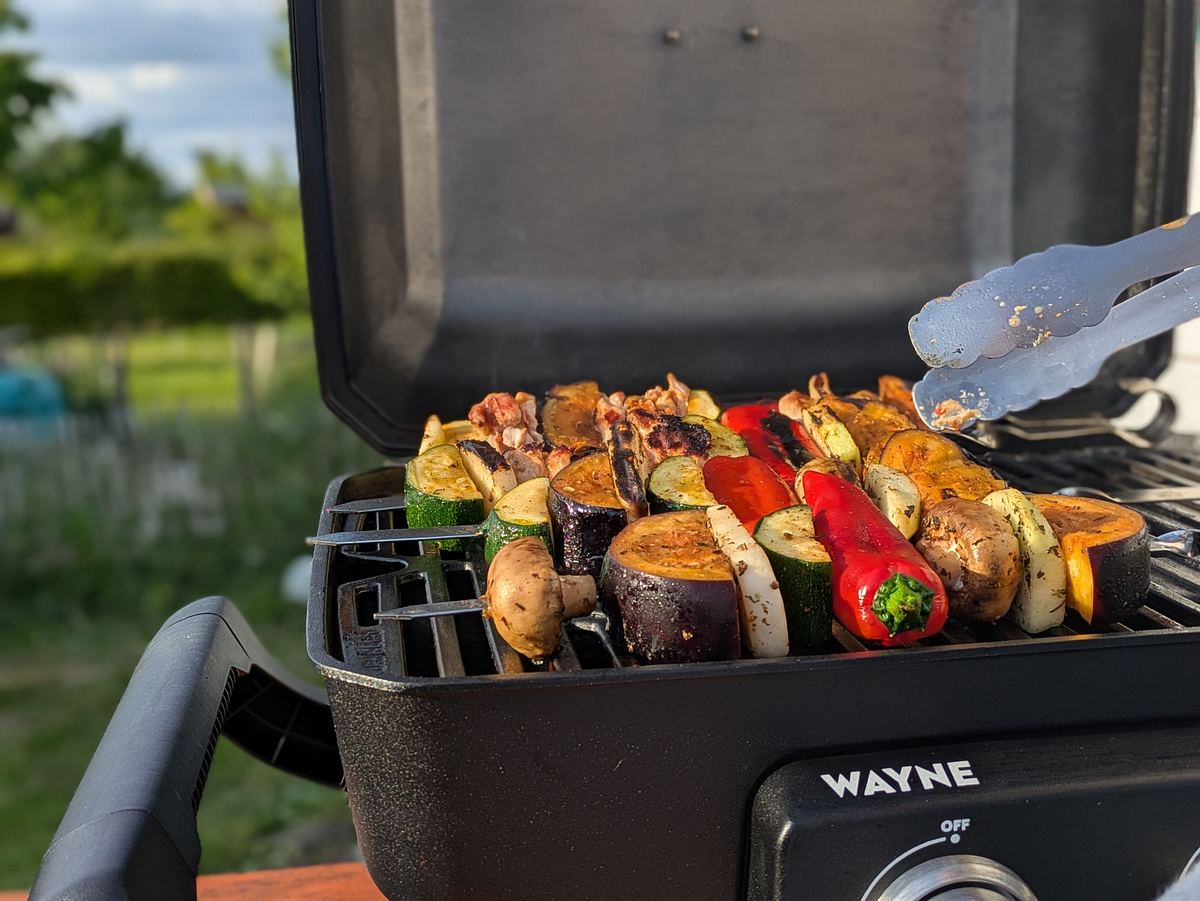 burnhard-wayne-gasgrill-gemuese-spiesse Ein geöffneter Burnhard Wayne Grill mit Spießen aus roter Paprika, Champignons, Aubergine und Zucchini, wobei eine Grillzange ein Stück anhebt; im Hintergrund sind Garten und Bäume sichtbar.