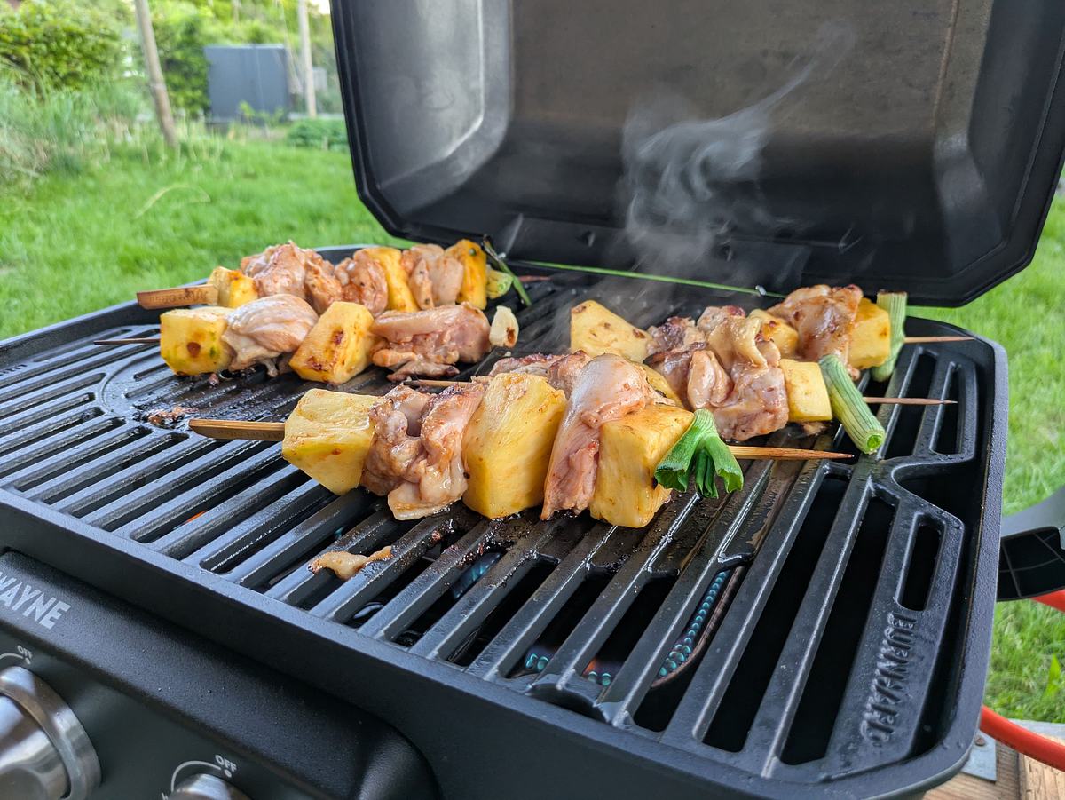 burnhard-gasgrill-wayne-spiesse Ein Burnhard Wayne Grill mit geöffnetem Deckel zeigt Spieße mit rohem Fleisch, Ananas und Frühlingszwiebeln, im Hintergrund ist Gras zu sehen.