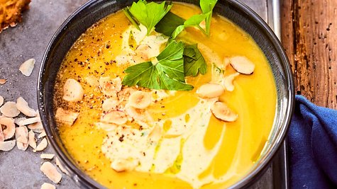 Gelbe Butternut-Kürbissuppe in einer dunklen Schale serviert mit Haselnusblättchen, Kräutern und Sahne garniert. - Foto: LECKER @ Bauer Media Group