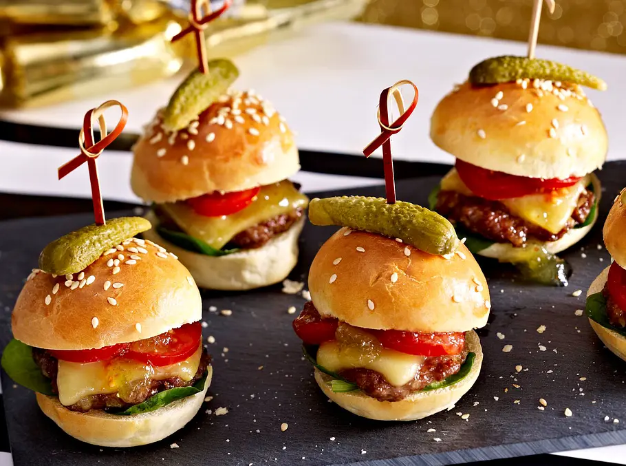 4 kleine Mini-Burger mit jeweils einem Cornichon am Spieß auf einer schwarzen Servierplatte serviert.
