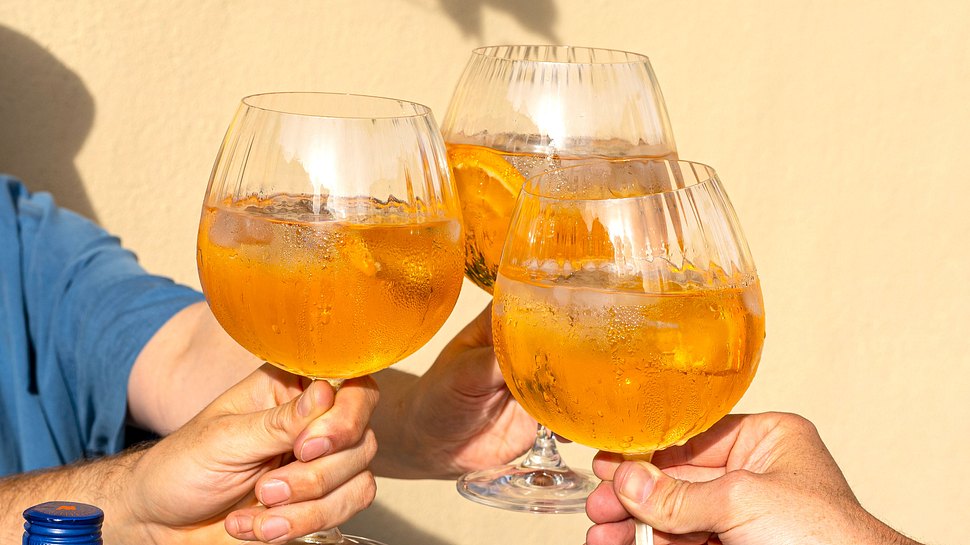 Drei bauchig geriffelte Gläser mit Canonita Spritz in Händen, beim Anstoßen. - Foto: Eggers & Franke