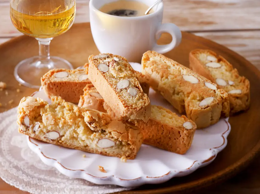 Goldbraune Cantuccini mit ganzen Mandeln liegen auf einem weißen Teller. Im Hintergrund ist eine gefüllte Espressotasse und ein Glas Vin Santo zu sehen.