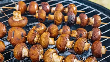  Mehrere Holzspieße mit braunen Champignons liegen nebeneinander auf einem Grillrost über glühenden Kohlen. - Foto: ShowHeroes für LECKER @ Bauer Media Group