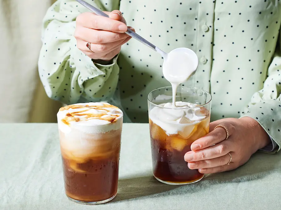 Zwei Gläser mit Cheesecake-Chai stehen im Vordergrund. Eine Hand gießt die Cheesecake-Creme als Topping in eines der Gläser