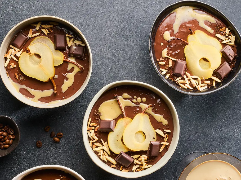 Vier Schalen mit cremigem Schoko-Kaffee-Pudding, jeweils mit pochierten Birnenscheiben, Schokoladenstücken und gehackten Mandeln garniert, dazu eine Schale mit Kaffeebohnen und ein Glas cremiger Kaffee auf dunkler Unterlage.