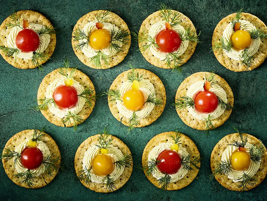 Runde Cracker mit einer Cremehaube, garniert mit roten und gelben Kirschtomaten sowie Dill auf einer dunklen Platte angerichtet.