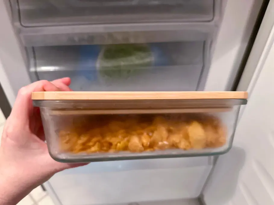 Eine Hand hält eine eiskalte Glasbox mit Cornflakes im Inneren, wobei im Hintergrund ein offener Gefrierschrank zu sehen ist