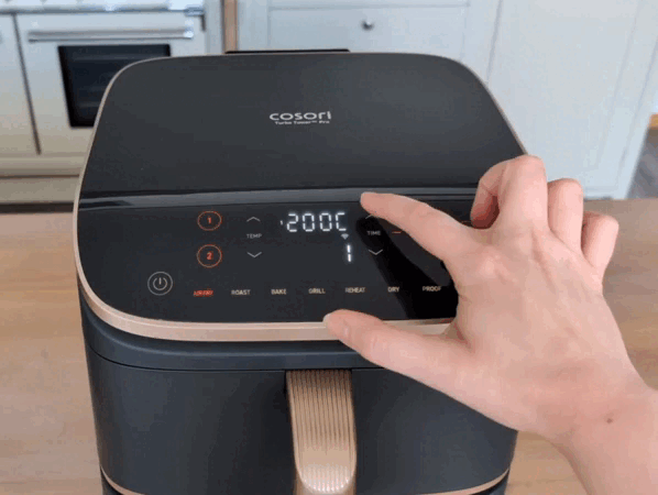 Mit dem Zeigefinger tippt sich die Testerin durch das Einstellungsmenü des Cosori Turbo Tower Pro Smart und wählt nacheinander Temperatur sowie Garzeit für beide Kochzonen aus.