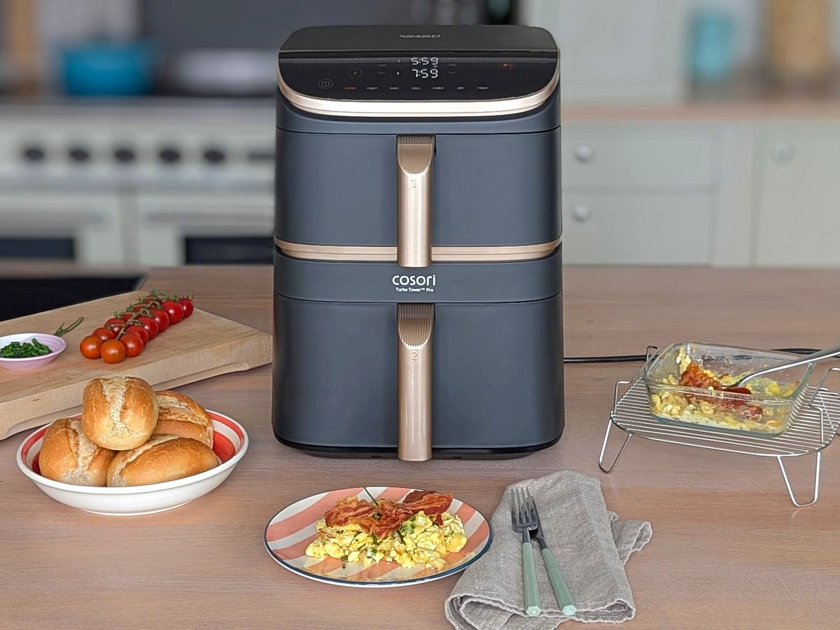 Der Cosori Turbo Tower Pro Smart steht auf einer Arbeitsfläche. Davor liegen Brötchen in einer Schale, Rührei mit Bacon auf einem Teller, Besteck und eine Glasform mit fertigem Rührei auf dem Gitterrost des Airfryers. Im Hintergrund liegen Schnittlauch und Tomaten auf einem Holzbrett.