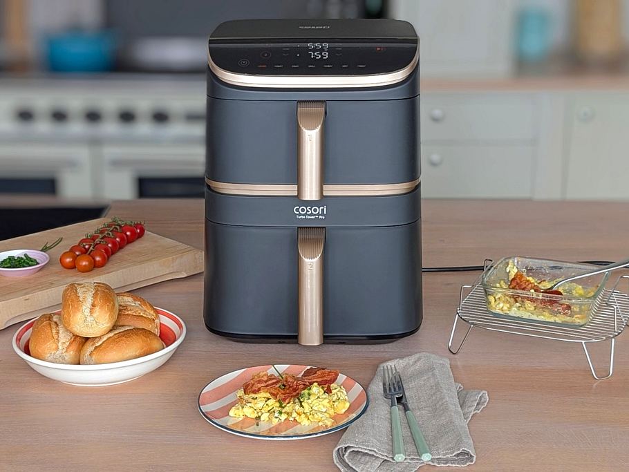 Der Cosori Turbo Tower Pro Smart steht auf einer Arbeitsfläche. Davor liegen Brötchen in einer Schale, Rührei mit Bacon auf einem Teller, Besteck und eine Glasform mit fertigem Rührei auf dem Gitterrost des Airfryers. Im Hintergrund liegen Schnittlauch und Tomaten auf einem Holzbrett.