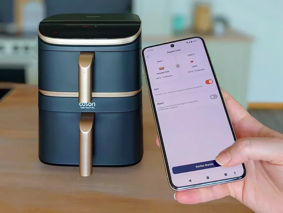 Im Hintergrund steht der Cosori Turbo Tower Pro Smart auf einer Arbeitsfläche in der Küche. Im Vordergrund hält eine Hand ein Smartphone, auf dem die VeSync-App läuft. Zu sehen ist ein eingestellter Kochvorgang im „Sync“-Modus mit Pommes in Zone 1 und Lachs in Zone 2. Der Daumen der Hand liegt auf dem Menüpunkt „Kochen starten“.