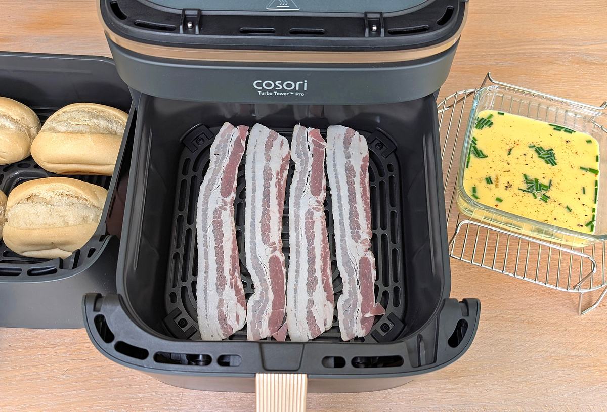 Brötchen, Bacon und Rühreimasse vor dem Garen Der Cosori Turbo Tower Pro Smart steht mit geöffneter unterer Schublade auf einer Arbeitsfläche. Darin liegen rohe Bacon-Scheiben. Links daneben steht die andere Schublade mit vier ungebackenen Brötchen. Rechts steht der Bratrost mit einer Glasschüssel darauf, die eine Rühreimasse enthält.