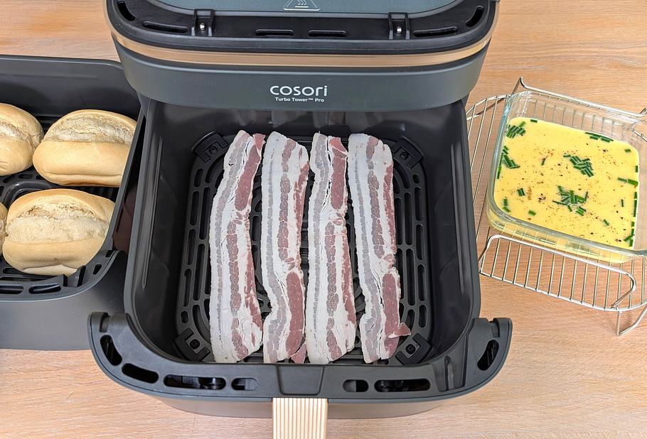 Der Cosori Turbo Tower Pro Smart steht mit geöffneter unterer Schublade auf einer Arbeitsfläche. Darin liegen rohe Bacon-Scheiben. Links daneben steht die andere Schublade mit vier ungebackenen Brötchen. Rechts steht der Bratrost mit einer Glasschüssel darauf, die eine Rühreimasse enthält.