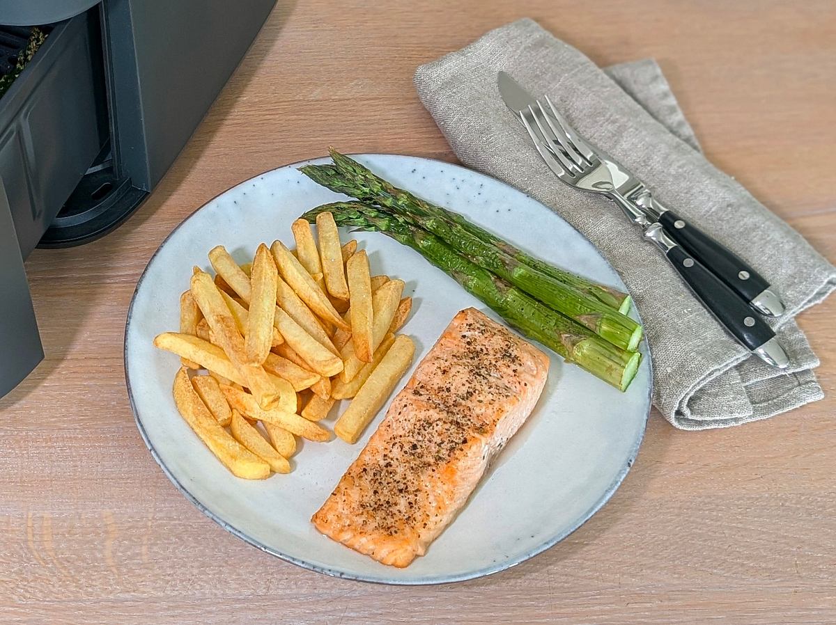 Lachs, Pommes und Spargel aus dem Cosori Turbo Tower Pro Smart. Rechts neben dem Cosori Turbo Tower Pro Smart steht ein Teller mit Pommes, Spargel und Lachsfilet. Wir sehen den Teller von oben. Rechts davon liegen Messer und Gabel auf einer Serviette.