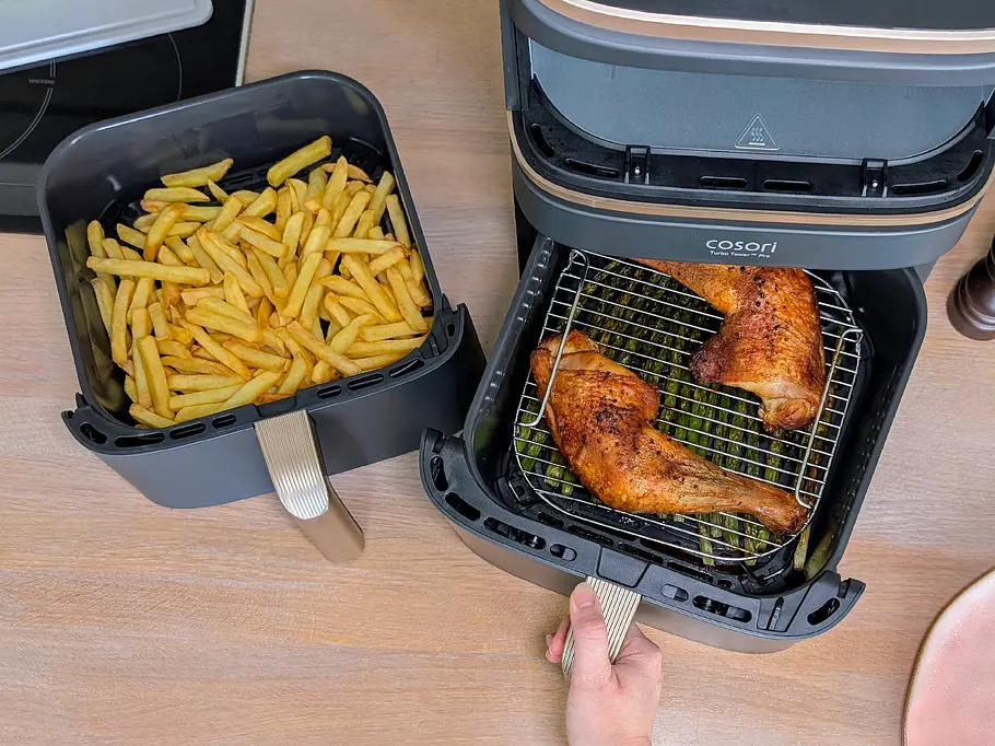 Blick von oben auf eine Holzarbeitsfläche. Auf ihr stehen der Cosori Turbo Tower Pro Smart, dessen oberes Fach leer ist, und seine beiden Schubladen: eine links neben ihm, befüllt mit goldgelben Pommes, die andere wird gerade mit einer Hand aus dem Airfryer gezogen. In dieser liegen zwei gebratene Hähnchenschenkel auf einem Gitterrost.