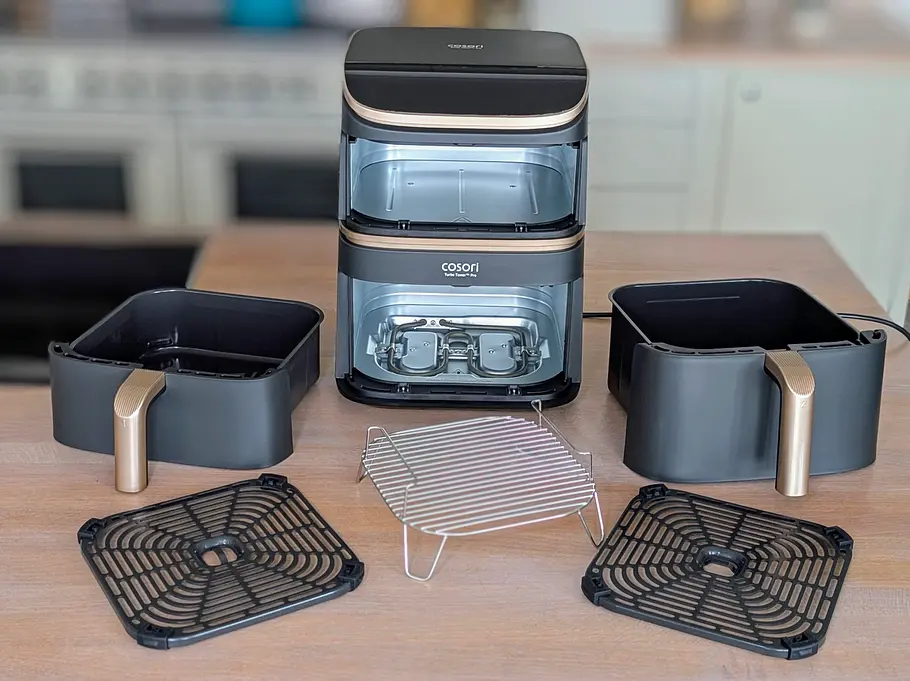 Der Cosori Turbo Tower Pro Smart steht in der Mitte einer Arbeitsfläche. Um ihn herum all seine Zubehörteile: die beiden unterschiedlich hohen Schubladen, zwei Knusperplatten, ein Bratrost.