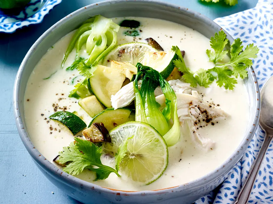 Cremige Kokos-Suppe mit Hähnchen Cremige, weiße Kokossuppe in einer Schale, darin Stücke von Hähnchen, Zucchini, Limettenscheiben und frischer Koriander. Die Suppe ist mit grobem Pfeffer bestreut und wird von einem Löffel und Serviette begleitet.