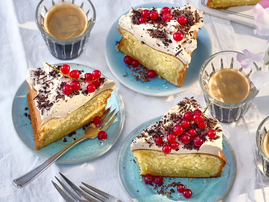 Stücke cremiger Eiscafé-Cheesecake auf hellblauen Tellern, mit Sahne, Schokoraspeln und roten Johannisbeeren garniert, dazu Espressotassen auf weißem Stoff.