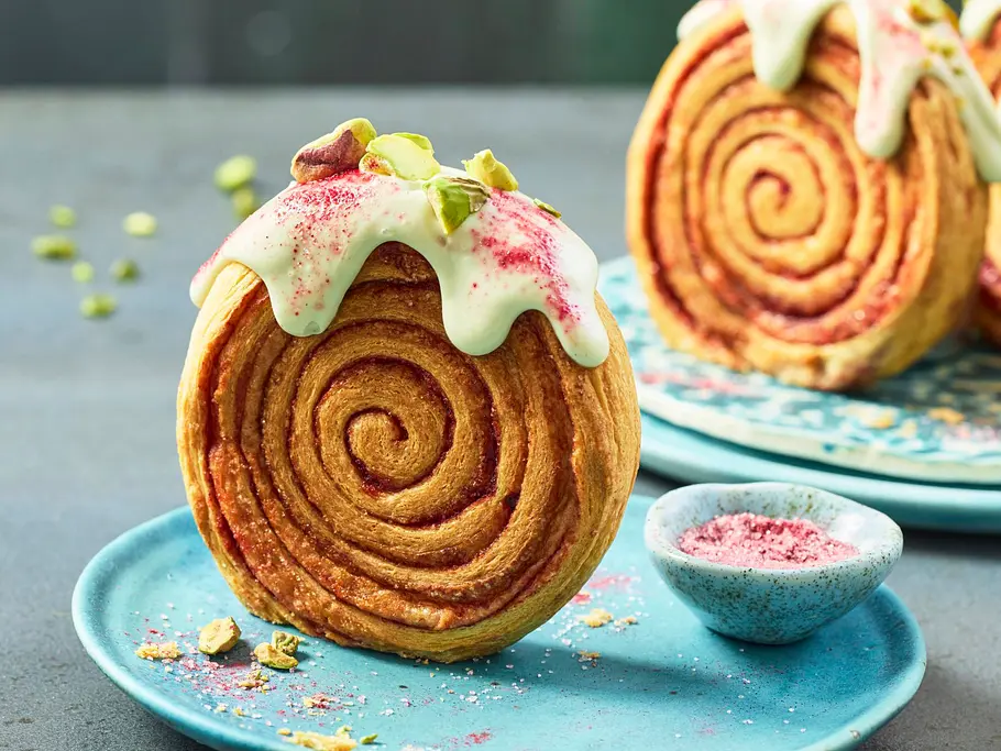 Croissant-Rollen mit Pistazien-Frosting auf blauem Teller, dekoriert mit gehackten Pistazien und pinkem Topping.