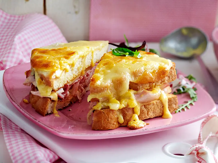 Croque Monsieur mit Hollandaise Rezept Auf einem rosa Teller liegt ein schräg angeschnittenes Monsieur-Croque-Sandwich, das außen goldbraun mit Käse überbacken und innen dick mit Kochschinken belegt ist.