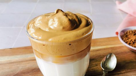 Ein Glas Dalgona Coffee mir karamellfarbener Cremehaube auf Milch. Daneben ein kleiner Löffel auf einem Holzbrett. - Foto: Birte Wloka / LECKER.de