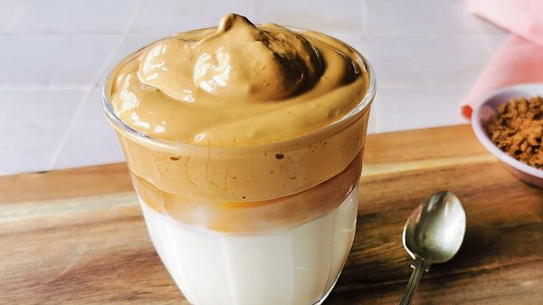 Ein Glas Dalgona Coffee mir karamellfarbener Cremehaube auf Milch. Daneben ein kleiner Löffel auf einem Holzbrett. - Foto: Birte Wloka / LECKER.de