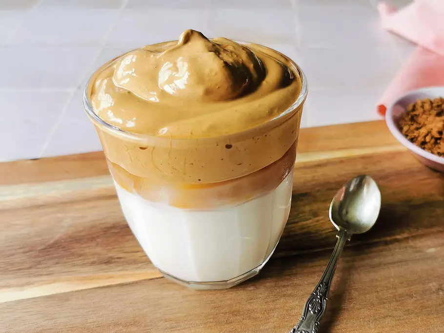 Dalgona Coffee Rezept Ein Glas Dalgona Coffee mir karamellfarbener Cremehaube auf Milch. Daneben ein kleiner Löffel auf einem Holzbrett.