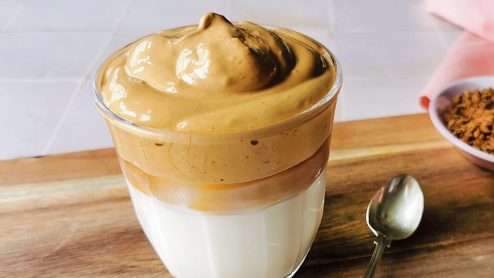 Ein Glas Dalgona Coffee mir karamellfarbener Cremehaube auf Milch. Daneben ein kleiner Löffel auf einem Holzbrett. - Foto: Birte Wloka / LECKER.de