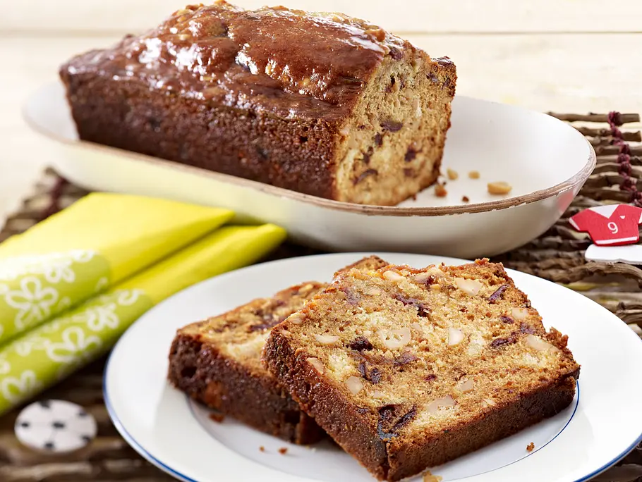 Saftiger Kastenkuchen mit sichtbaren Dattelstücken und gehackten Erdnüssen, in Scheiben auf einem weißen Teller angerichtet, der restliche Laib auf einer ovalen Platte im Hintergrund.