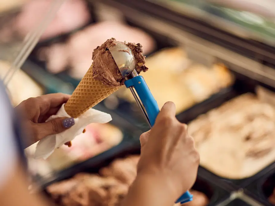 Nahaufnahme eines Schokoladeneises in einer Waffel, das von einer Eisverkäuferin mit einer Kugel vorbereitet wird.