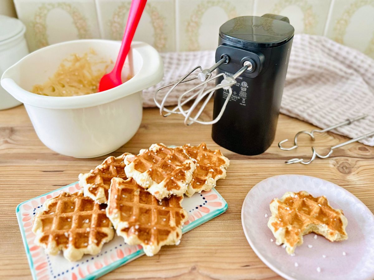 Auf einer hölzernen Arbeitsfläche steht ein kabelloses Handrührgerät mit eingesetzten Quirlen neben einer Schüssel mit Waffelteig und einer Servierplatte mit frischen  Lütticher Waffeln