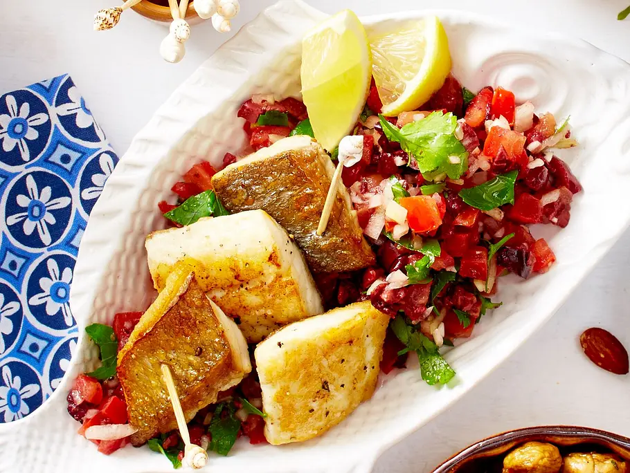 Goldbraun gebratene Doradenfilets auf einem Sommersalat aus gehacktem Gemüse, angerichtet auf einer weißen Fischschale, daneben liegen Zitronenspalten.