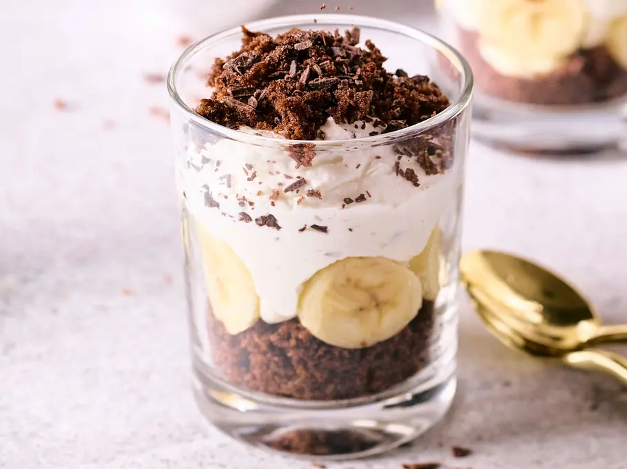 Dreischichtiger Maulwurf-Hügel im Glas mit Schokoladenkuchen, Bananenscheiben und Sahnecreme, getoppt mit Schokoraspeln, daneben goldene Löffel auf heller Oberfläche.