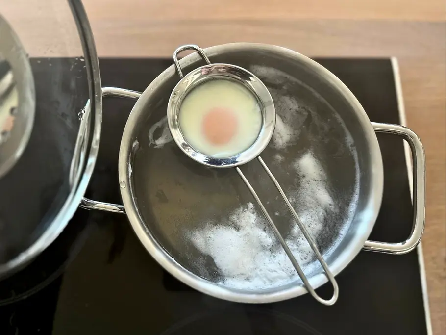 Eier pochieren im Sieb In einem Topf mit kochendem Wasser ist ein kleines Küchensieb mit einem halb gestockten Ei eingehängt.