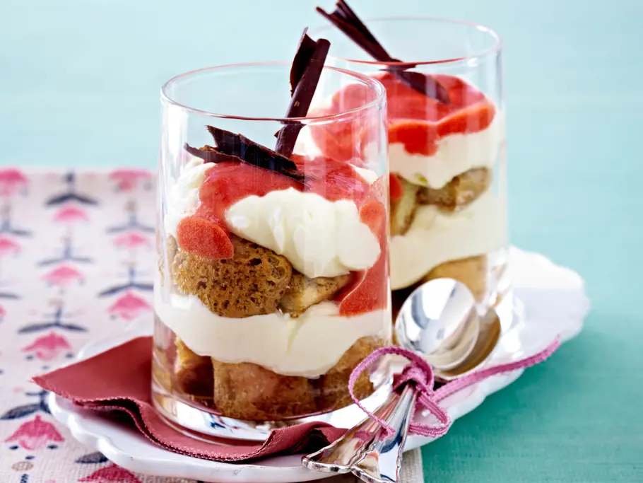 Eierlikör-Tiramisu Rezept Zwei Dessertgläser mit geschichteten Löffelbiskuits, die in Espresso braun getränkt sind, mit weißer Creme und roter Erdbeersoße getoppt sind.