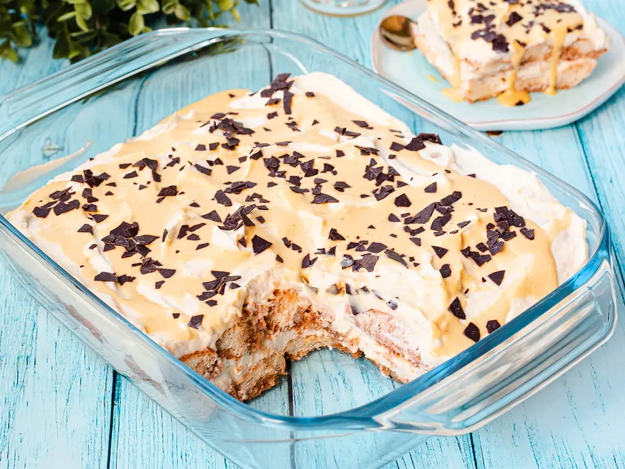 Cremiges Oster-Tiramusu Rezept Das Ostertiramisu befindet sich in einer gläsernen Auflaufform. Eine Portion ist bereits entnommen, sodass man die Schichtung aus Biskuit und weißer Sahne-Mascarpone-Creme sieht. Das Tiramisu ist mit hellgelbem Eierlikör und Schokoraspel garniert. Das entnommene Portion befindet sich auf einem Teller im Hintergrund.