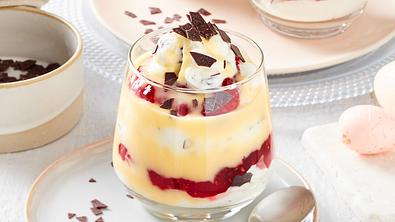 Im Glas werden weiße Quarkcreme, Himbeerkonfitüre und Eierlikör geschichtet. Garniert wird das Glas mit Eierlikörtraum mit Schokoraspel. Das Glas steht auf einen Teller, auf dem außerdem ein Dessertlöffel liegt. Im Hintergrund sind ein weiteres Glas und hellrosa Eier zu erkennen. - Foto: LECKER @ Bauer Media Group