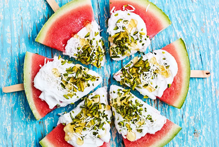 Eis am Stiel „One in a Melon“ Wassermelonenstücke mit Holzstäbchen, Kokoscreme, gehackten Pistazien und Kokosraspeln auf blauem Untergrund angerichtet.