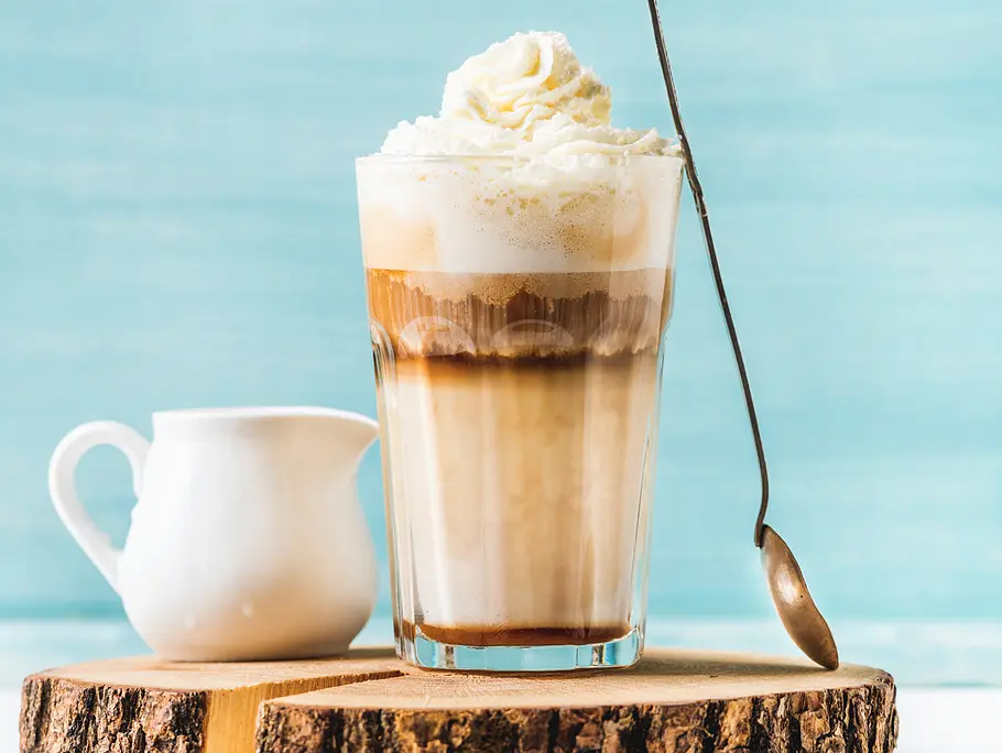 Ein hohes Glas mit Milch, Kaffee und einer Eiskugel gefüllt. Getoppt mit Schlagsahne. Am Glas lehnt ein langer Löffel