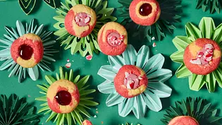 Runde Engelsaugen-Plätzchen mit rot-gelbem Teig, gefüllt mit roter Marmelade oder rosa Zuckerguss und zerkleinerten Candy Canes, auf grünen Papier-Sternunterlagen angerichtet. Eine Hand hält ein Plätzchen. - Foto: LECKER @ Bauer Media Group