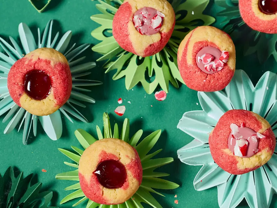 Runde Engelsaugen-Plätzchen mit rot-gelbem Teig, gefüllt mit roter Marmelade oder rosa Zuckerguss und zerkleinerten Candy Canes, auf grünen Papier-Sternunterlagen angerichtet. Eine Hand hält ein Plätzchen.