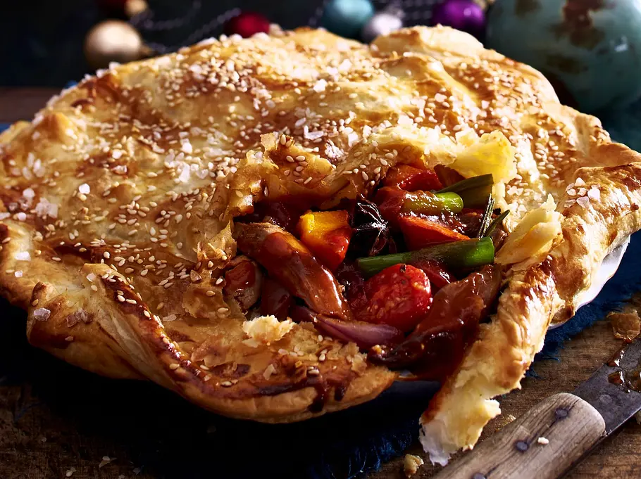 Christmas-Pie mit goldbrauner Blätterteighaube, gefüllt mit buntem Gemüse und zartem Fleisch in herzhafter Soße, serviert in einer runden Backform auf einem rustikalen Holzbrett.