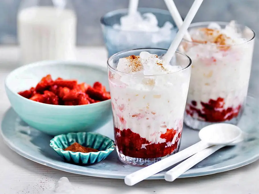 Auf einem Tabeltt stehen zwei Gläser mit rotem Erdbeerpüree, das von cremiger Buttermilch und Crushed Ice getoppt wird, neben einer Schale mit frischen halbierten Erdbeeren und Eiswürfeln.