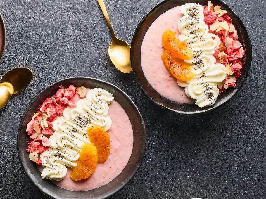 Vier Schüsseln mit cremigem, rosa Erdbeer-Pudding, getoppt mit Blutorangenfilets, Joghurtcreme, gehackten Nüssen und gefriergetrockneten Erdbeerstückchen, serviert auf einem dunklen Untergrund. Daneben ein Teller mit Blutorangenscheiben und Nüssen.
